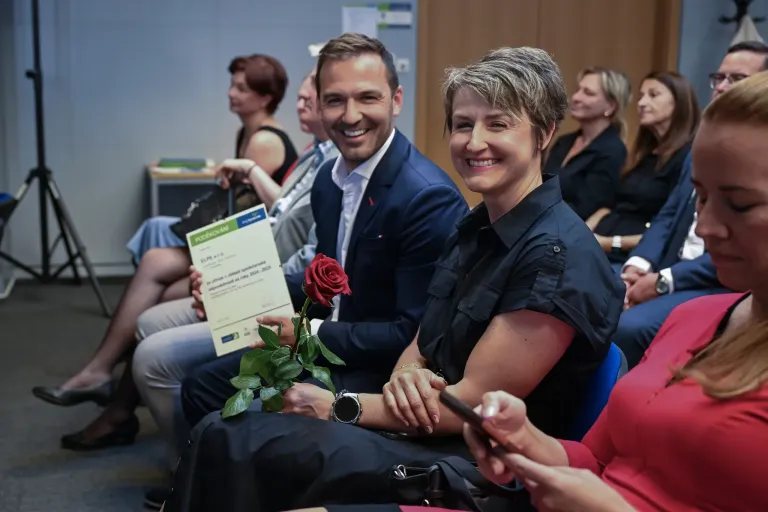 Skupina lidí sedící na společenské akci s certifikátem a růží v ruce