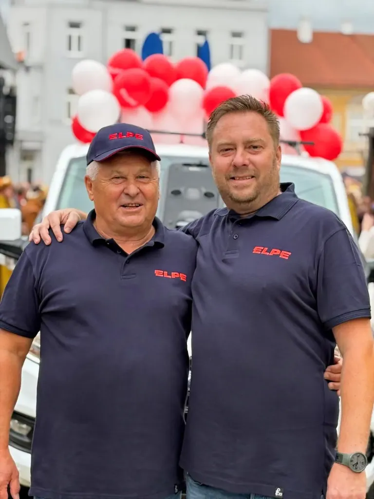 Dva muži v tričkách s logem ELPE objímající se na městské slavnosti s balónky
