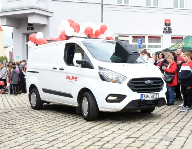 Bílý dodávkový vůz ELPE s reproduktorem a balónky projíždí mezi lidmi na náměstí