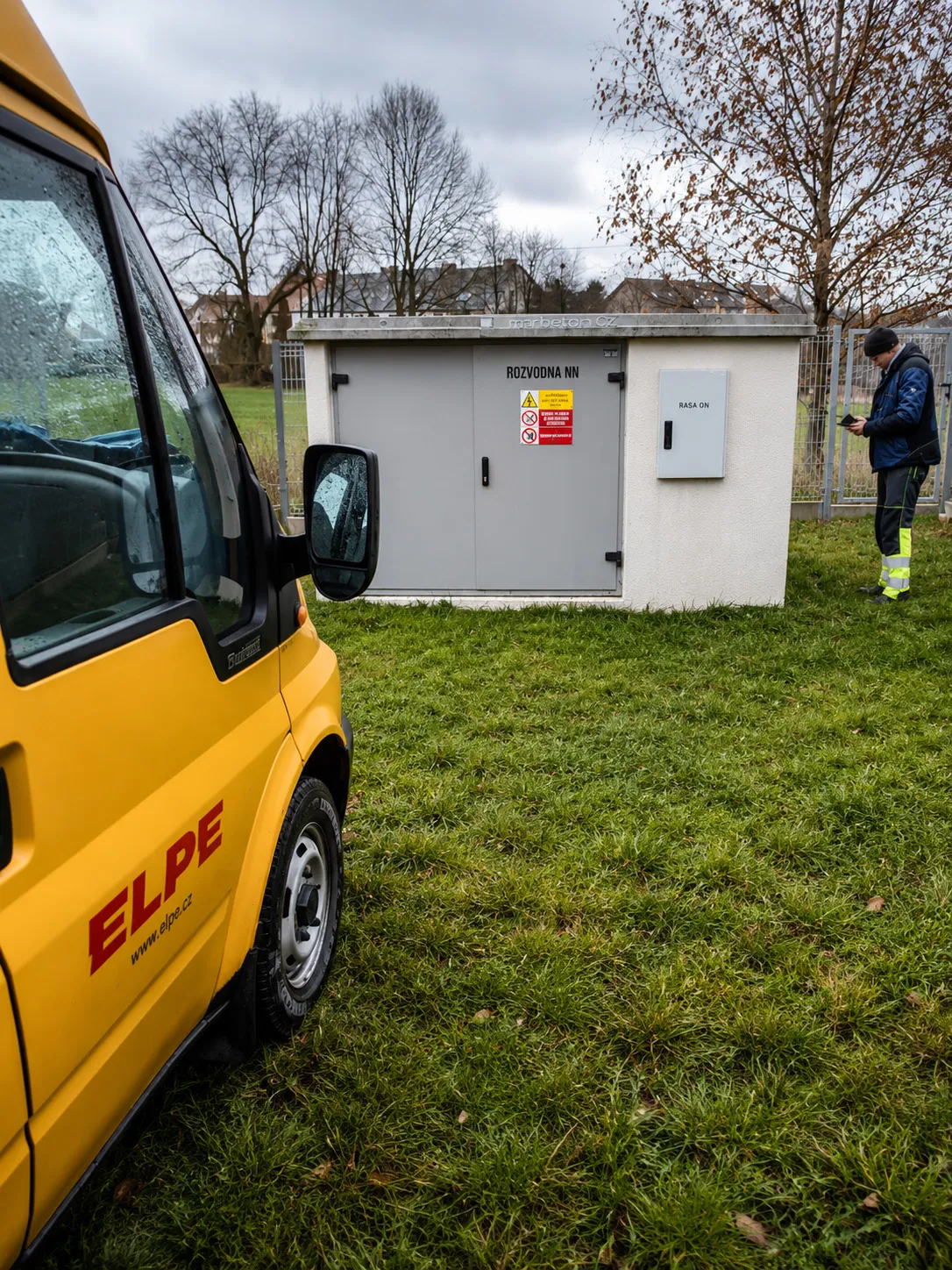 Nízkonapěťová trafostanice s žlutým vozem ELPE a pracovníkem kontrolujícím zařízení na trávníku